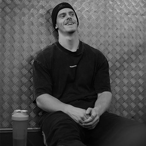 Titan Dylan zit ontspannen tegen een metalen wand in de gym met een shakebeker naast zich.